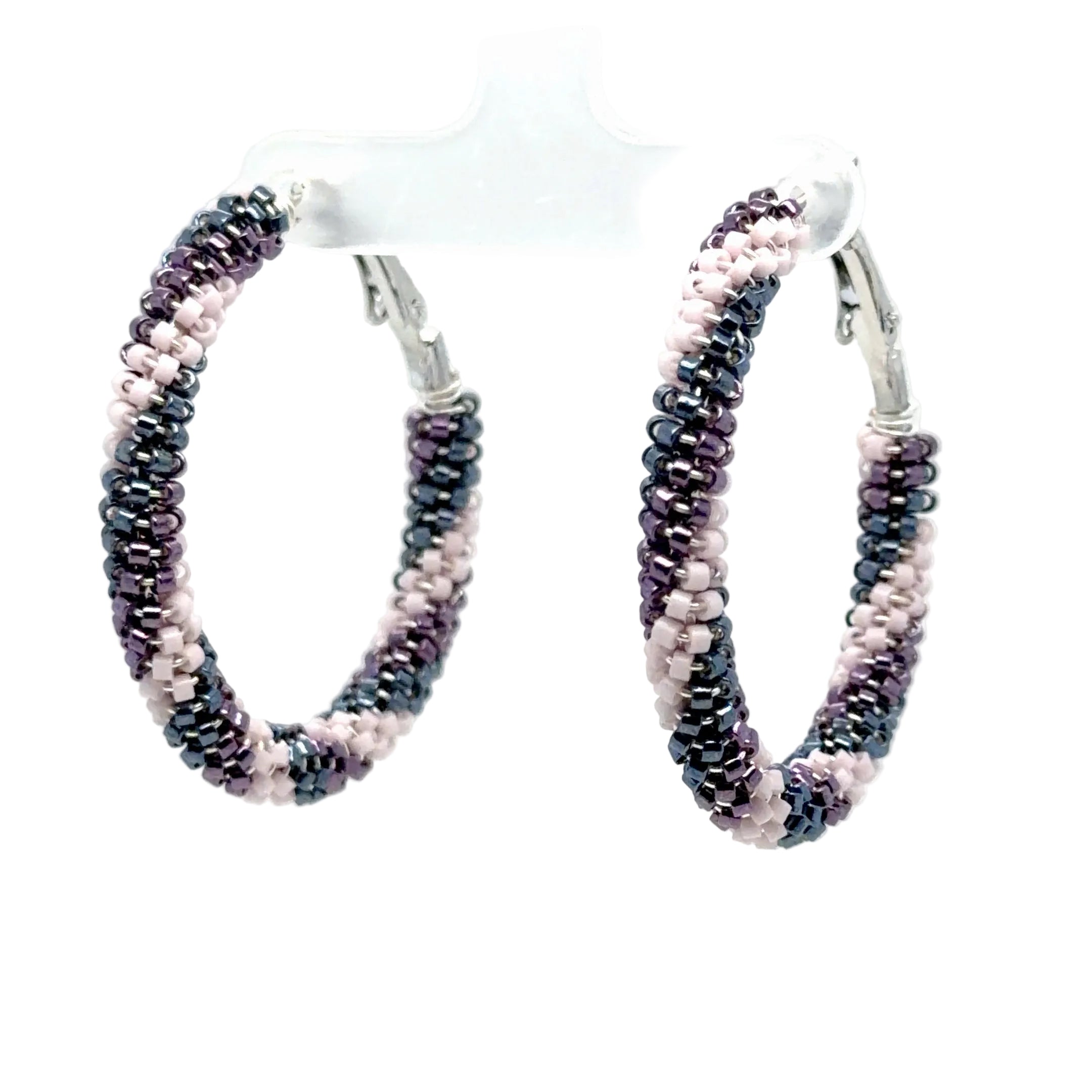 A pair of medium hoop earrings made from small seed beads in alternating dark metallic gray and light pink, hanging from silver-toned fishhook clasps on a clear display stand against a white background.