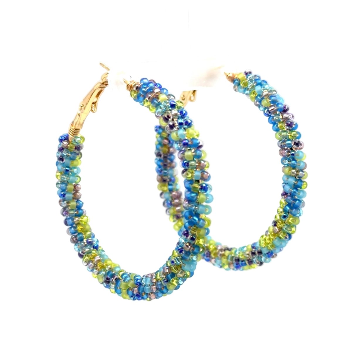 A pair of large hoop earrings made from small round beads in blue, green, and purple tones, with gold-tone clasp hardware, displayed on a white stand against a white background.