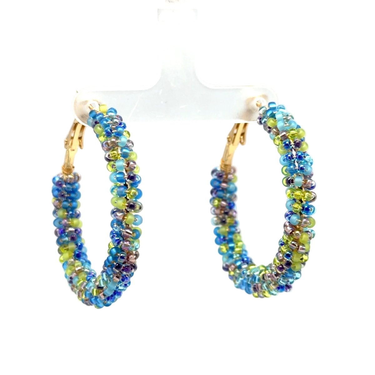 A pair of beaded hoop earrings with gold-tone fishhook backs, featuring strands of iridescent blue, green, and purple beads, displayed on a clear plastic stand against a white background.