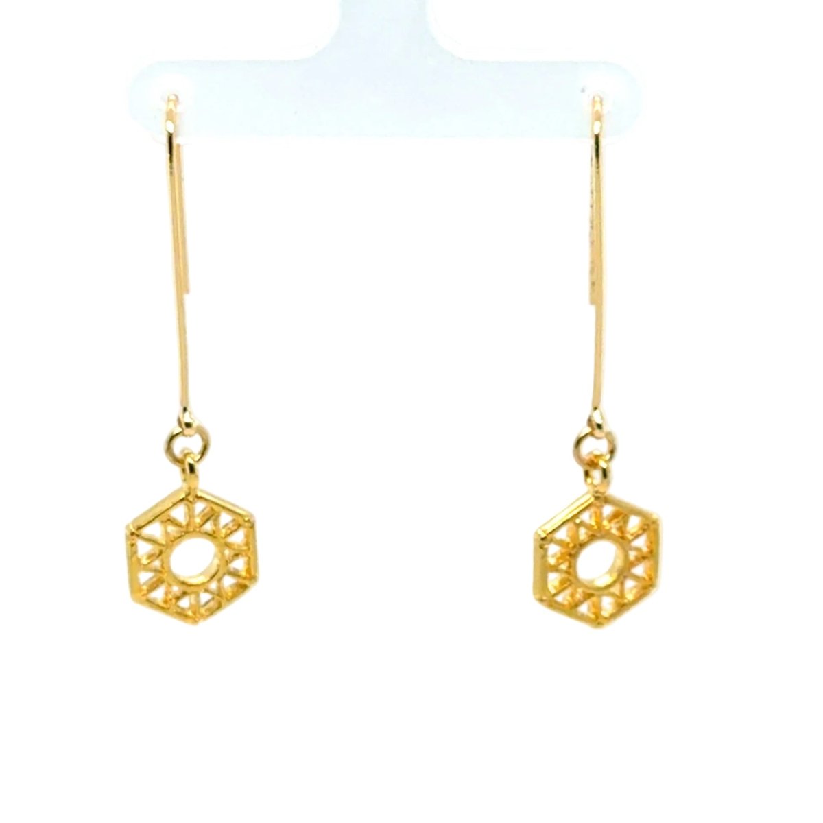 A pair of gold geometric dangle earrings with a hexagonal openwork design, hanging from thin gold hooks on a white display stand.