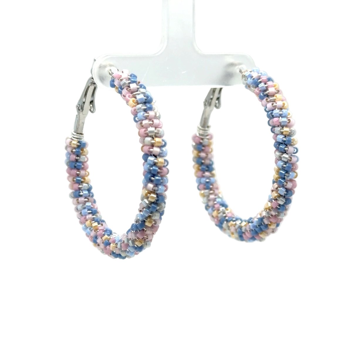 A pair of large hoop earrings made of tightly woven pastel beads in blue, pink, and gold, hanging from silver-toned clips on a clear display stand against a white background.